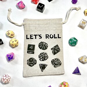Op de afbeelding: Een beige trekkoordtas met de tekst "LET'S ROLL" erop gedrukt. De tas is omringd door verschillende kleurrijke dobbelstenen, waaronder een groene d20, een paarse d12, een witte d8 en een rode d4.