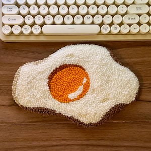 May include: A handmade, egg-shaped rug with a white, textured surface and a brown border. The yolk is a vibrant orange, and the rug is placed on a wooden surface. A cream-colored keyboard is in the background.