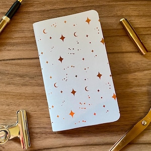 Op de afbeelding: Een wit notitieboek met een herhalend patroon van metallic koperkleurige sterren, maansikkels en kleine stippen. Het notitieboek is omringd door een gouden vulpen, een gouden pendop, een zilveren metalen klem en een gouden schaar.