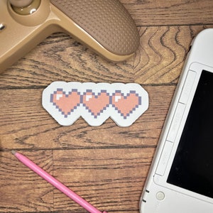 May include: Three pixelated hearts in a row on a white background. The hearts are orange and are on a wooden surface. A pink stylus and a brown video game controller are also in the image.