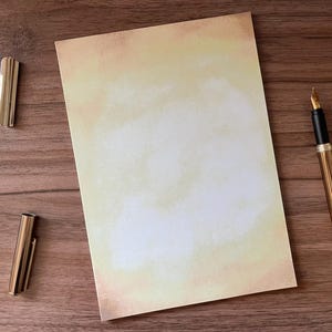 Könnte beinhalten: Ein Schreibwarenset mit cremefarbenem Papier mit Aquarelleffekt, einem goldenen Füllfederhalter, einer goldenen Büroklammer und einer goldenen Stiftkappe. Die Artikel sind auf einer Holzoberfläche angeordnet.