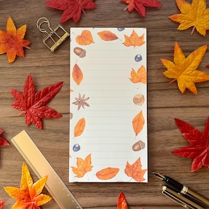 May include: A lined notepad featuring autumn leaf, acorn, and berry illustrations. The notepad is surrounded by colourful artificial maple leaves, a gold binder clip, a gold ruler, and a pen on a wooden surface.