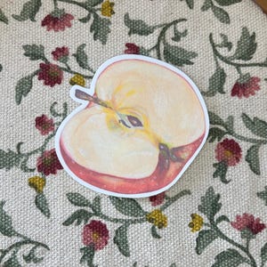 Puede incluir: Una pegatina de manzana roja con un centro blanco, que muestra una sección transversal de la manzana. La pegatina está sobre un fondo blanco con un patrón floral.