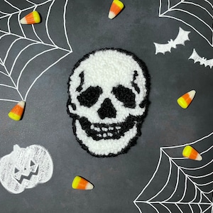 May include: A white and black yarn skull with a black background. The skull is surrounded by candy corn and spiderwebs.