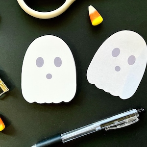 May include: Two white ghost-shaped sticky notes with gray oval eyes and a small gray circular mouth. The scene includes a silver metal binder clip, a black retractable ballpoint pen, and four pieces of orange, yellow, and white candy corn.