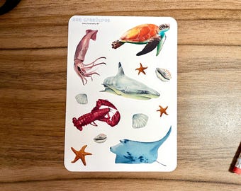 May include: A sheet of stickers featuring various sea creatures, including a squid, a shark, a lobster, a sea turtle, and a manta ray. The stickers are on a white background with a few starfish and seashells.