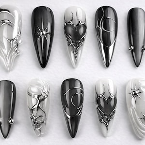 May include: A collection of ten long, pointed artificial nails. The nails feature a variety of designs, including solid black, white, and gradient colors. Some nails have silver metallic embellishments, such as starbursts, hearts, and abstract shapes.