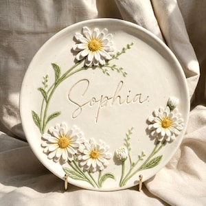 May include: A round, off-white ceramic decorative plate featuring the name Sophia embossed in the centre. The plate is adorned with raised, three-dimensional white daisy flowers with yellow centres and green stems and leaves arranged around the text.