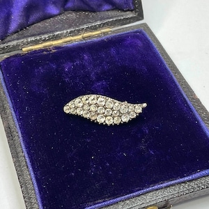 May include: A vintage silver-toned brooch in the shape of a leaf, encrusted with numerous small, clear gemstones. The brooch rests on a deep blue velvet lining within a dark, antique box, suggesting a jewelry or antique item.