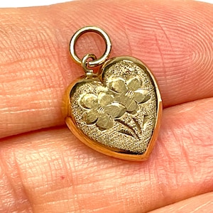 May include: A gold heart-shaped pendant featuring a textured surface with an engraved floral design. The pendant includes a small circular jump ring at the top for attachment. The metal has a polished gold finish with contrasting matte details.