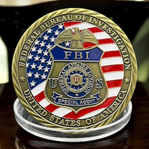 Puede incluir: Una moneda conmemorativa dorada con la inscripción "FEDERAL BUREAU OF INVESTIGATION" y "UNITED STATES OF AMERICA." La moneda presenta un diseño azul, blanco y rojo con un águila y las letras "FBI." Las palabras "SPECIAL AGENT" también son visibles.