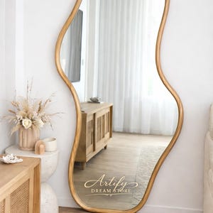 May include: A large, gold-framed, free-standing mirror with an organic, wavy shape. The mirror leans against a white wall, reflecting a room with wooden furniture and a neutral color palette. The text "Artify Dream Store" is visible on the mirror.