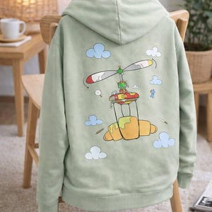 Może przedstawiać: Szałwiowa zielona bluza z kapturem z fantazyjną grafiką na plecach. Projekt przedstawia rogalik z śmigłem helikoptera, otoczony chmurami i małą postacią. Bluza z kapturem jest prezentowana na drewnianym krześle.