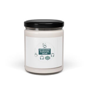 May include: A white candle in a glass jar with a black lid. The label features the text "Introvert Survival Gear" with icons of a house, headphones, a mug, and a blanket. The candle is designed to create a relaxing atmosphere.