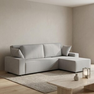 Light gray modular sectional couch with chaise lounge, photographed in a modern living room on a textured neutral area rug. The sofa features ribbed corduroy fabric, broad armrests, deep seating, two oversized back cushions, and two matching side pillows for added comfort. A low stone-style coffee table with minimal decor sits in the foreground, emphasizing the sofa’s contemporary silhouette, soft upholstery texture, and inviting multifunctional layout.