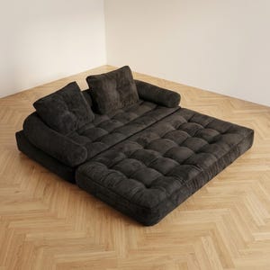 Dark charcoal grey corduroy futon sofa bed fully unfolded flat into its bed configuration, placed in a bright room corner on a light herringbone parquet floor. The two-piece tufted mattress surface is spread wide, with the two back cushions resting at the head end and the rounded side armrests visible on each side. The wide ribbed corduroy texture and deep button tufting are clearly visible across the entire sleeping surface, demonstrating the spacious double bed size of this convertible sofa.