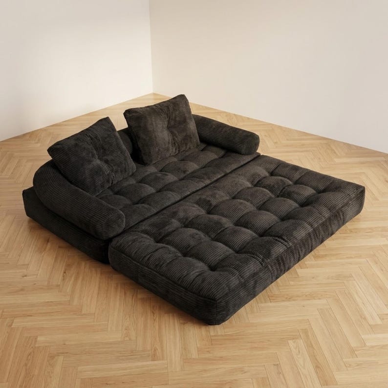 Dark charcoal grey corduroy futon sofa bed fully unfolded flat into its bed configuration, placed in a bright room corner on a light herringbone parquet floor. The two-piece tufted mattress surface is spread wide, with the two back cushions resting at the head end and the rounded side armrests visible on each side. The wide ribbed corduroy texture and deep button tufting are clearly visible across the entire sleeping surface, demonstrating the spacious double bed size of this convertible sofa.