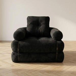 Deep black corduroy single-seater futon armchair in sofa configuration displayed against a plain white wall on a light herringbone parquet floor, photographed from a straight front-facing angle. The oversized plush chair features one large square tufted back cushion, a deeply tufted seat, and two large rounded armrests, all upholstered in wide-ribbed corduroy fabric. Dark metal buckle straps are visible on each side. A bold convertible armchair bed perfect for modern minimalist interiors.