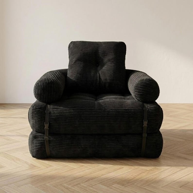 Deep black corduroy single-seater futon armchair in sofa configuration displayed against a plain white wall on a light herringbone parquet floor, photographed from a straight front-facing angle. The oversized plush chair features one large square tufted back cushion, a deeply tufted seat, and two large rounded armrests, all upholstered in wide-ribbed corduroy fabric. Dark metal buckle straps are visible on each side. A bold convertible armchair bed perfect for modern minimalist interiors.