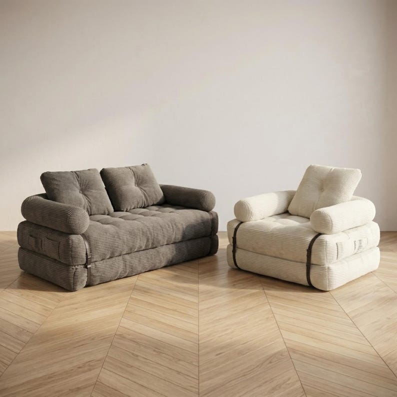 Two corduroy futon sofa beds displayed side by side in sofa configuration against a plain white wall on a light herringbone parquet floor, showcasing two available color options. On the left is a larger two-seater version in warm taupe grey with two large back cushions and a deeply tufted seat. On the right is a smaller single-seater version in cream off-white with one back cushion. Both feature wide-ribbed corduroy upholstery, rounded armrests, and dark buckle straps on the sides.