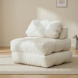 Beige convertible floor sofa chair displayed upright with a thick layered base and oversized tufted back cushions in a bright, minimalist room. The ribbed corduroy fabric texture, rounded edges, and pillow-like construction are clearly visible, placed on a neutral rug over light wood flooring. Soft sunlight creates gentle shadows that define the channels and stitching. Clean wall, simple styling, and centered framing highlight the compact single-seat form and cozy lounge profile.