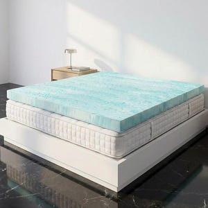 Può includere: Un materasso con un topper in schiuma blu chiaro e bianco. Il materasso è bianco e poggia su una struttura letto a piattaforma bianca. Una piccola lampada e un comodino sono sullo sfondo.