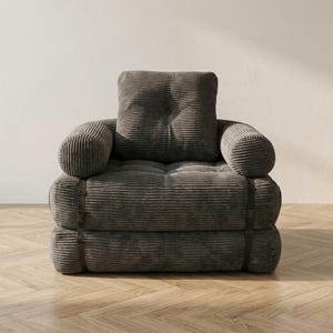 Gray corduroy single-seater futon armchair in sofa configuration displayed against a plain white wall on a light herringbone parquet floor, photographed from a straight front-facing angle. The oversized plush chair features one large square tufted back cushion, a deeply tufted seat, and two large rounded armrests, all upholstered in wide-ribbed corduroy fabric. Dark metal buckle straps are visible on each side. A bold convertible armchair bed perfect for modern minimalist interiors.