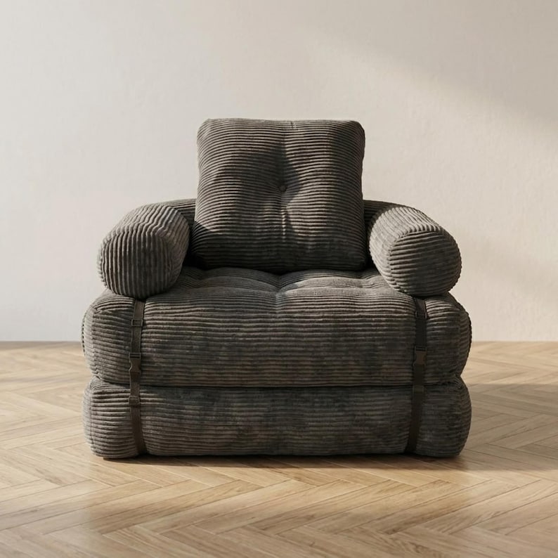 Gray corduroy single-seater futon armchair in sofa configuration displayed against a plain white wall on a light herringbone parquet floor, photographed from a straight front-facing angle. The oversized plush chair features one large square tufted back cushion, a deeply tufted seat, and two large rounded armrests, all upholstered in wide-ribbed corduroy fabric. Dark metal buckle straps are visible on each side. A bold convertible armchair bed perfect for modern minimalist interiors.