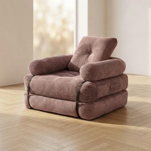 Dusty rose pink corduroy single-seater futon armchair in sofa configuration, placed in a bright sunlit room near a large window on a light herringbone parquet floor. The plush oversized chair features one large square tufted back cushion, a deeply tufted seat, and two rounded armrests, all covered in wide-ribbed corduroy fabric. Dark metal buckle straps are visible on the front and sides. A cozy and stylish convertible armchair that folds flat into a single bed for small apartments.