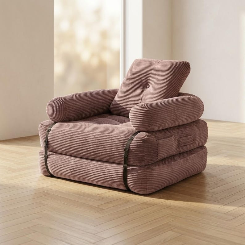 Dusty rose pink corduroy single-seater futon armchair in sofa configuration, placed in a bright sunlit room near a large window on a light herringbone parquet floor. The plush oversized chair features one large square tufted back cushion, a deeply tufted seat, and two rounded armrests, all covered in wide-ribbed corduroy fabric. Dark metal buckle straps are visible on the front and sides. A cozy and stylish convertible armchair that folds flat into a single bed for small apartments.