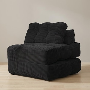 Black modular floor sofa chair in ribbed corduroy, photographed upright on a pale wood floor against a smooth neutral wall in a simple studio-like setting. The low, chunky base and plush tufted back cushions create a deep lounge-ready seat with rounded padded sides and visible channel stitching. Soft, even lighting shows the velvety corduroy ribs and clean seams. Minimal styling keeps attention on the rich black color, compact footprint, and versatile use as a floor couch or reading chair.