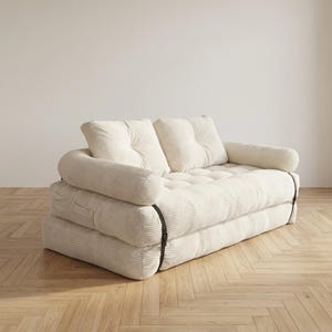Cream off-white corduroy fabric two-seater futon sofa bed displayed in sofa configuration against a plain white wall on a light herringbone parquet floor. The plush tufted seat cushion and two large back cushions are upholstered in a wide-ribbed corduroy texture, with soft rounded armrests on each side. Dark metal buckle straps are visible on the sides, securing the folded mattress layers. A stylish and versatile convertible sofa bed for small living spaces and minimalist modern home interiors.