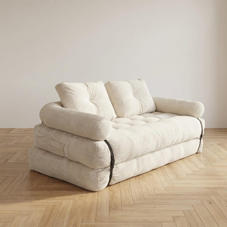 Cream off-white corduroy fabric two-seater futon sofa bed displayed in sofa configuration against a plain white wall on a light herringbone parquet floor. The plush tufted seat cushion and two large back cushions are upholstered in a wide-ribbed corduroy texture, with soft rounded armrests on each side. Dark metal buckle straps are visible on the sides, securing the folded mattress layers. A stylish and versatile convertible sofa bed for small living spaces and minimalist modern home interiors.