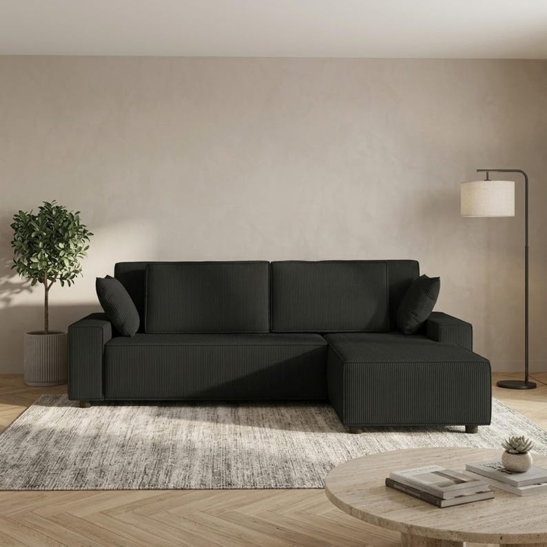 Black modular sectional sofa with chaise in ribbed corduroy fabric, displayed in a warm contemporary living room with a textured area rug and light wood flooring. The sofa features a low-profile silhouette, wide square arms, deep cushioned seating, two large back cushions, and matching accent pillows. A tall floor lamp, indoor plant, and round coffee table complete the scene, emphasizing the couch’s bold modern look, multifunctional design, and comfortable lounge-ready proportions.