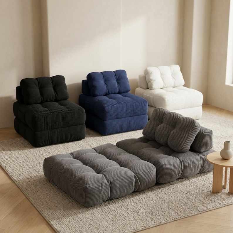Four-piece modular corduroy floor sofa set displayed in a minimalist living room: a dark gray chaise lounge unfolded on a textured rug in the foreground, with three single-seat sofa modules behind it in black, navy blue, and cream. Soft natural light, light wood flooring, and neutral walls highlight the plush ribbed fabric texture and stackable design.