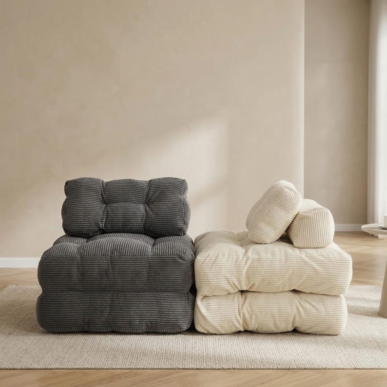 Two modular corduroy floor sofas placed side by side on a neutral rug: one dark gray unit in an upright chair configuration and one cream/beige unit stacked with matching cushions. The image highlights contrasting color options, plush ribbed fabric, and low-profile lounge seating design. Minimalist living room background with warm light and clean styling keeps focus on the product shape and texture.