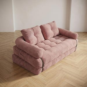 Dusty rose pink corduroy fabric two-seater futon sofa bed photographed from a slightly elevated angle in sofa configuration, placed on a light herringbone parquet floor in a bright minimalist room. The plush tufted seat and two large back cushions are covered in a wide-ribbed corduroy texture, with soft rounded armrests framing each side. The layered base structure is visible on the sides. A charming and space-saving convertible sofa bed ideal for small apartments and cozy living room setups.