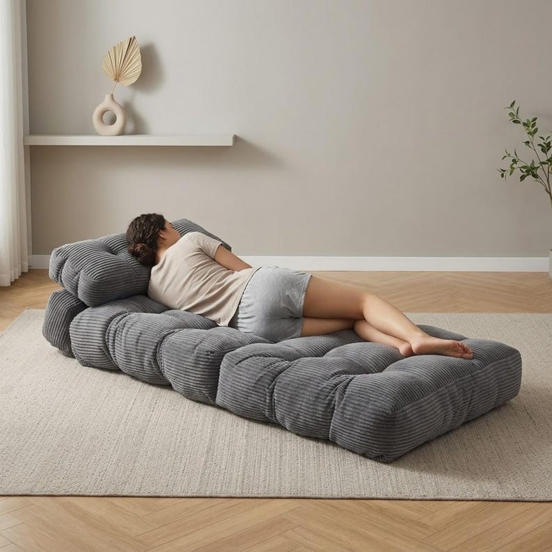 Gray corduroy foldable floor sofa mattress shown fully unfolded in a clean living room setting. A person lies on their side along the length of the sofa, viewed from behind, demonstrating lounging comfort and full-body support. The ribbed plush cushions form a low chaise-style bed on a large neutral rug with light wood flooring and a simple modern wall décor for scale.