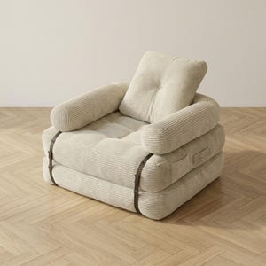 Cream off-white corduroy single-seater futon armchair in sofa configuration displayed against a plain white wall on a light herringbone parquet floor. The plush oversized chair features one large square tufted back cushion, a deeply tufted seat, and two rounded armrests, all covered in wide-ribbed corduroy fabric. Dark metal buckle straps are clearly visible on the front and sides. A soft and elegant convertible armchair bed ideal for minimalist Scandinavian and modern home interiors.