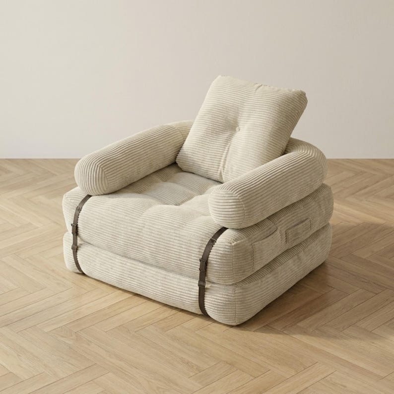 Cream off-white corduroy single-seater futon armchair in sofa configuration displayed against a plain white wall on a light herringbone parquet floor. The plush oversized chair features one large square tufted back cushion, a deeply tufted seat, and two rounded armrests, all covered in wide-ribbed corduroy fabric. Dark metal buckle straps are clearly visible on the front and sides. A soft and elegant convertible armchair bed ideal for minimalist Scandinavian and modern home interiors.