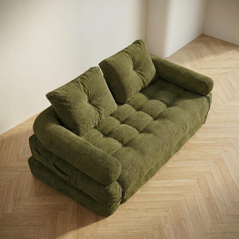 Olive green corduroy fabric two-seater futon sofa bed photographed from a slightly elevated angle in sofa configuration, placed in a bright minimalist room corner on a light herringbone parquet floor. The plush tufted seat and two large back cushions are upholstered in a wide-ribbed corduroy texture in a warm earthy green tone, with soft rounded armrests on each side. The layered base structure is visible from this angle. A bold and stylish convertible sofa bed for retro-inspired modern.