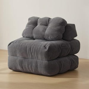 Dark gray modular corduroy floor chair in upright sofa configuration, photographed in a minimalist interior. Thick ribbed cushions create a deep seat with a plush backrest and side pillow support, showing the stacked design and soft texture. Neutral wall backdrop and light wood floor emphasize the compact footprint and cozy lounge style, suitable for reading corners, dorms, or small spaces.