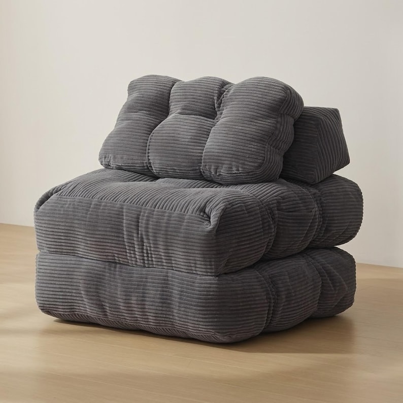Dark gray modular corduroy floor chair in upright sofa configuration, photographed in a minimalist interior. Thick ribbed cushions create a deep seat with a plush backrest and side pillow support, showing the stacked design and soft texture. Neutral wall backdrop and light wood floor emphasize the compact footprint and cozy lounge style, suitable for reading corners, dorms, or small spaces.