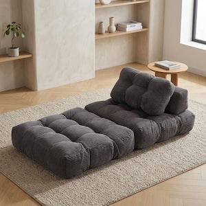 May include: A modular gray corduroy floor sofa with a tufted design. The sofa consists of a rectangular base and a backrest with two cushions. The sofa is placed on a neutral-colored rug in a room with light-colored walls and wooden floors.
