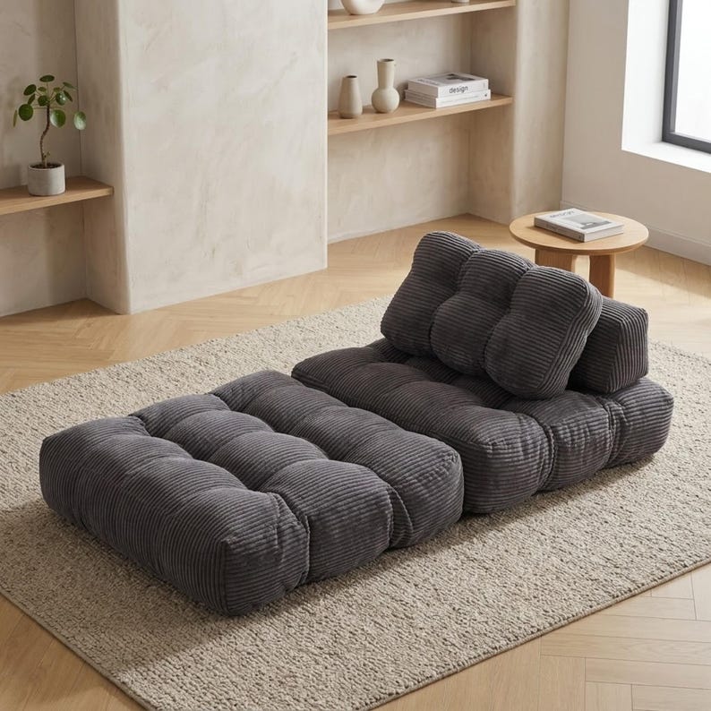 May include: A modular gray corduroy floor sofa with a tufted design. The sofa consists of a rectangular base and a backrest with two cushions. The sofa is placed on a neutral-colored rug in a room with light-colored walls and wooden floors.