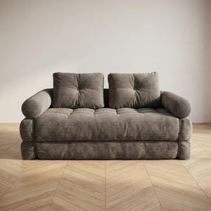 Warm taupe grey corduroy fabric two-seater futon sofa bed displayed in sofa configuration against a plain white wall on a light herringbone parquet floor, photographed from a straight front-facing angle. The deep tufted seat and two large square back cushions are upholstered in a wide-ribbed corduroy texture, with soft rounded armrests on each side. The thick layered base adds a cozy floor-level aesthetic. A versatile and stylish convertible sofa bed perfect for modern minimalist living rooms.