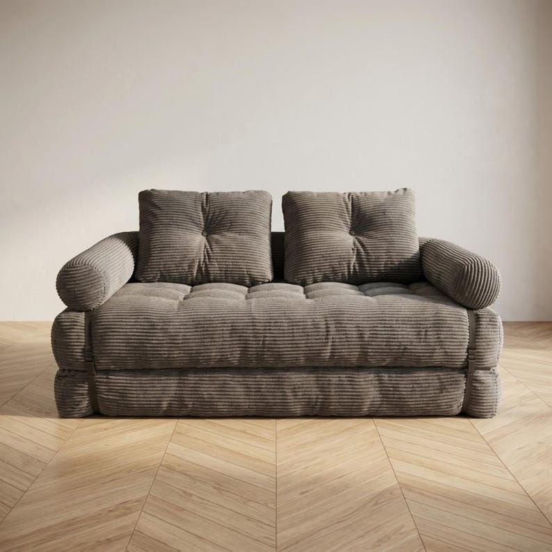 Warm taupe grey corduroy fabric two-seater futon sofa bed displayed in sofa configuration against a plain white wall on a light herringbone parquet floor, photographed from a straight front-facing angle. The deep tufted seat and two large square back cushions are upholstered in a wide-ribbed corduroy texture, with soft rounded armrests on each side. The thick layered base adds a cozy floor-level aesthetic. A versatile and stylish convertible sofa bed perfect for modern minimalist living rooms.