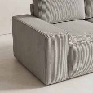 Close-up detail of a light gray corduroy sectional sofa arm, highlighting the wide boxy armrest, vertical ribbed upholstery, and crisp tailored edges. The image showcases the fabric’s soft texture, structured shape, and clean stitching, with part of the seat cushion visible beside the arm. Set against a smooth neutral floor and softly lit background, this detail photo emphasizes the sofa’s modern craftsmanship, durable finish, and elegant minimalist design for contemporary interiors.