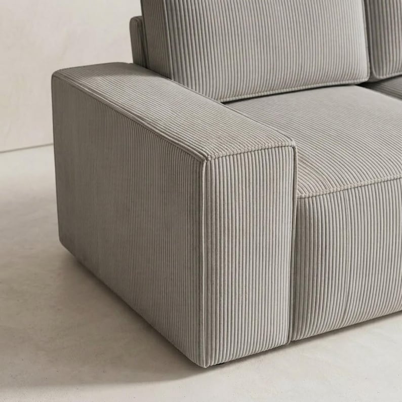 Close-up detail of a light gray corduroy sectional sofa arm, highlighting the wide boxy armrest, vertical ribbed upholstery, and crisp tailored edges. The image showcases the fabric’s soft texture, structured shape, and clean stitching, with part of the seat cushion visible beside the arm. Set against a smooth neutral floor and softly lit background, this detail photo emphasizes the sofa’s modern craftsmanship, durable finish, and elegant minimalist design for contemporary interiors.
