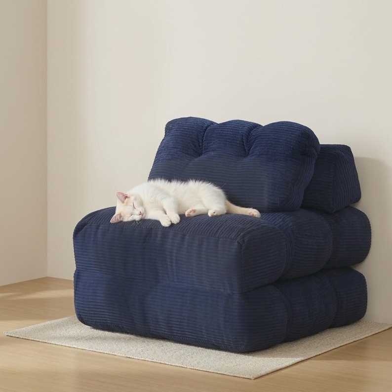 Navy blue modular floor sofa chair made from plush ribbed corduroy, shown upright in a minimalist living room corner on a light woven rug and pale wood floor. A small white cat is curled up sleeping on the wide cushioned seat, showing scale. The sofa features deep channel tufting, a thick puffy backrest with two pillows, rounded padded sides, and a low-profile stackable base. Neutral beige wall and soft natural daylight keep focus on texture, color, and cozy lounge use.